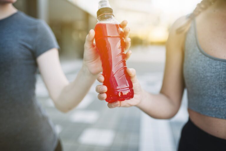 Two women exchanging a red electrolyte drink bottle outdoors, emphasizing hydration and energy replenishment.