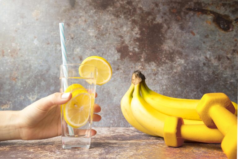 Glass of lemon water with bananas and yellow dumbbells in the background, symbolizing hydration and electrolyte-rich nutrition