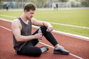Male athlete sitting on a running track, holding a water bottle to rehydrate after exercise.