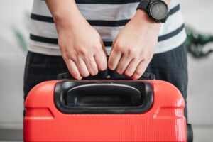 Close-up of a person holding the handle of a red suitcase, wearing a striped shirt and a black watch.
