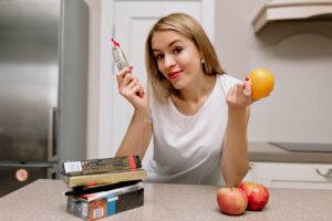 Young woman in a kitchen holding an orange and a packaged food item, with a stack of packaged foods and apples on the counter, symbolizing dietary choices and insulin resistance awareness.