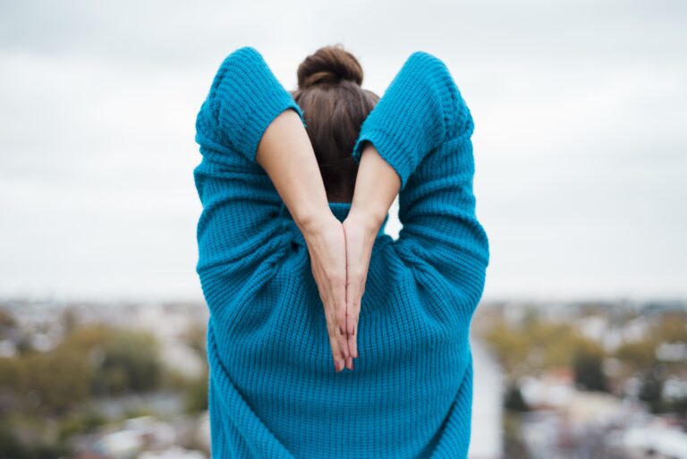 Woman stretching her arms overhead while wearing a blue sweater outdoors.